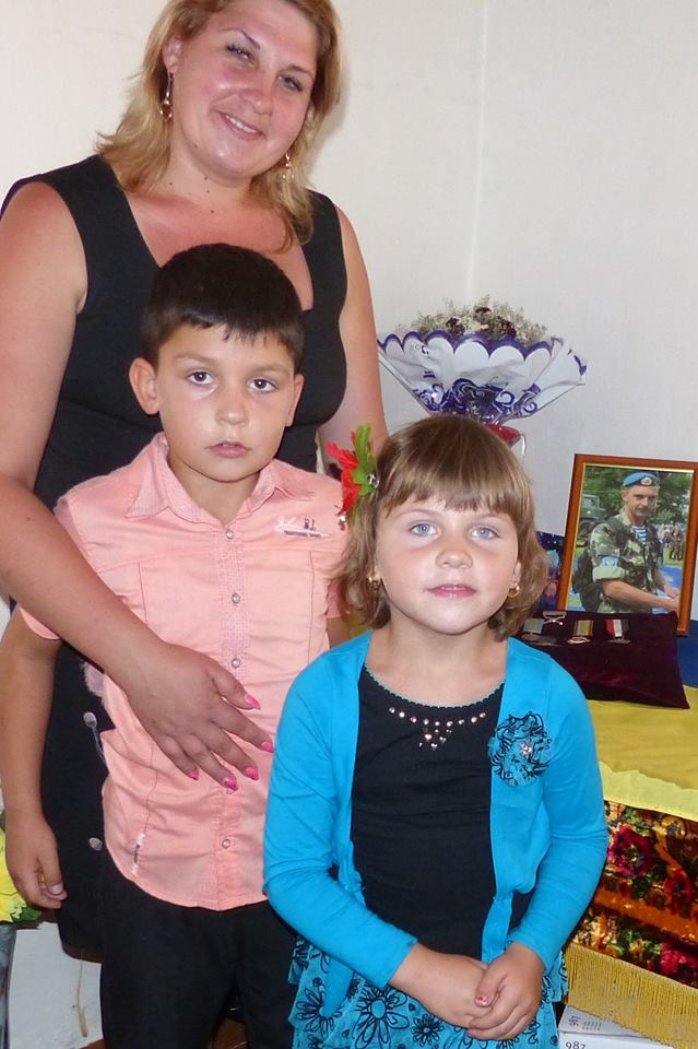 Maryna with her two children standing at a memorial to her husband Oleksandr, including a few pictures and his commendation for bravery.