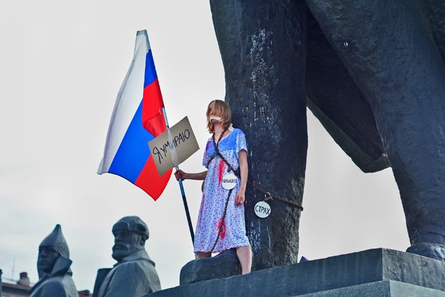 A woman dressed as Russia in a blood-stained outfit and holding a national flag chained herself to a Lenin statue in Novosibirsk, Russia to protest deteriorating human rights and increasing poverty in the country. The large sign attached to the flag says: "I Am Dying." Three smaller signs hanging off the chain crisscrossing her body say: "Police," "Fear" and "Censorship." August 22, 2017 (Image: Alyona Martynova / Sib.fm)