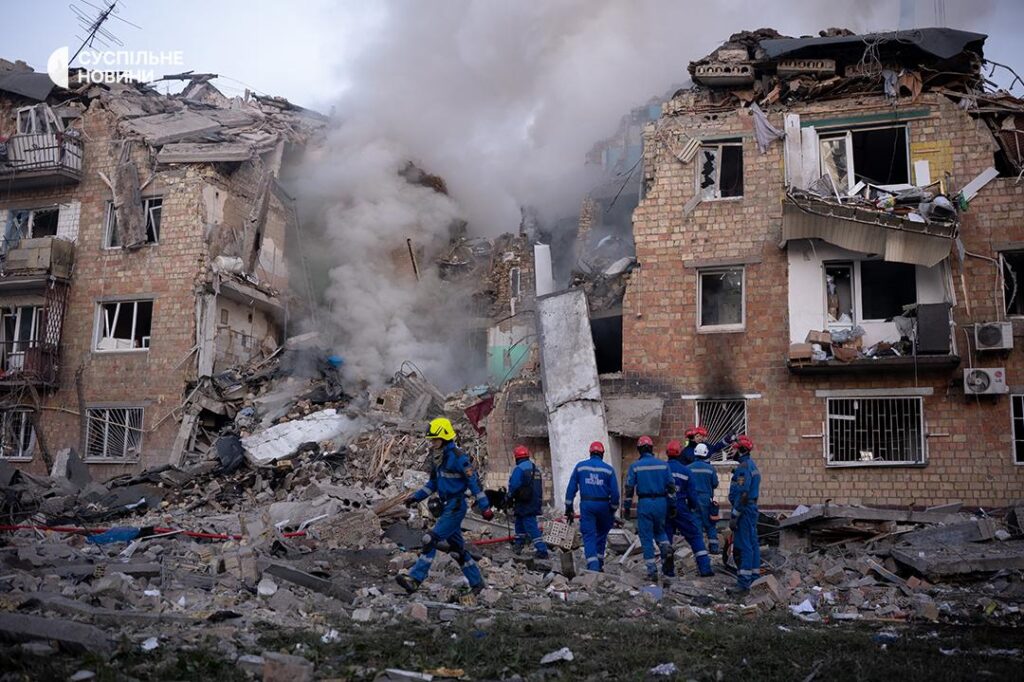 Emergency workers in blue uniforms search through debris and rubble of severely damaged multi-story residential buildings in Kyiv, Ukraine, with destroyed walls and broken windows, smoke visible in background