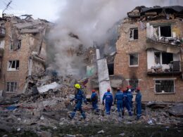 Emergency workers in blue uniforms search through debris and rubble of severely damaged multi-story residential buildings in Kyiv, Ukraine, with destroyed walls and broken windows, smoke visible in background