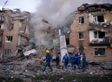 Emergency workers in blue uniforms search through debris and rubble of severely damaged multi-story residential buildings in Kyiv, Ukraine, with destroyed walls and broken windows, smoke visible in background