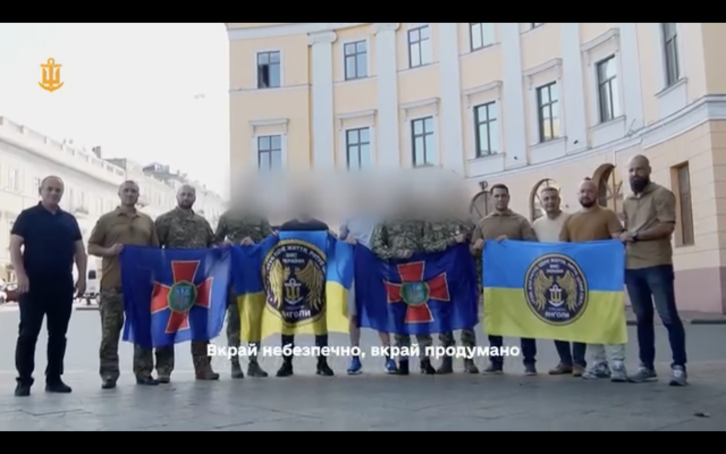Four Ukrainian soldiers hold National Guard and naval flags following their evacuation by special forces from a hospital on Russian-occupied territory where they had been hiding since 2022. Their faces are blurred for security reasons.