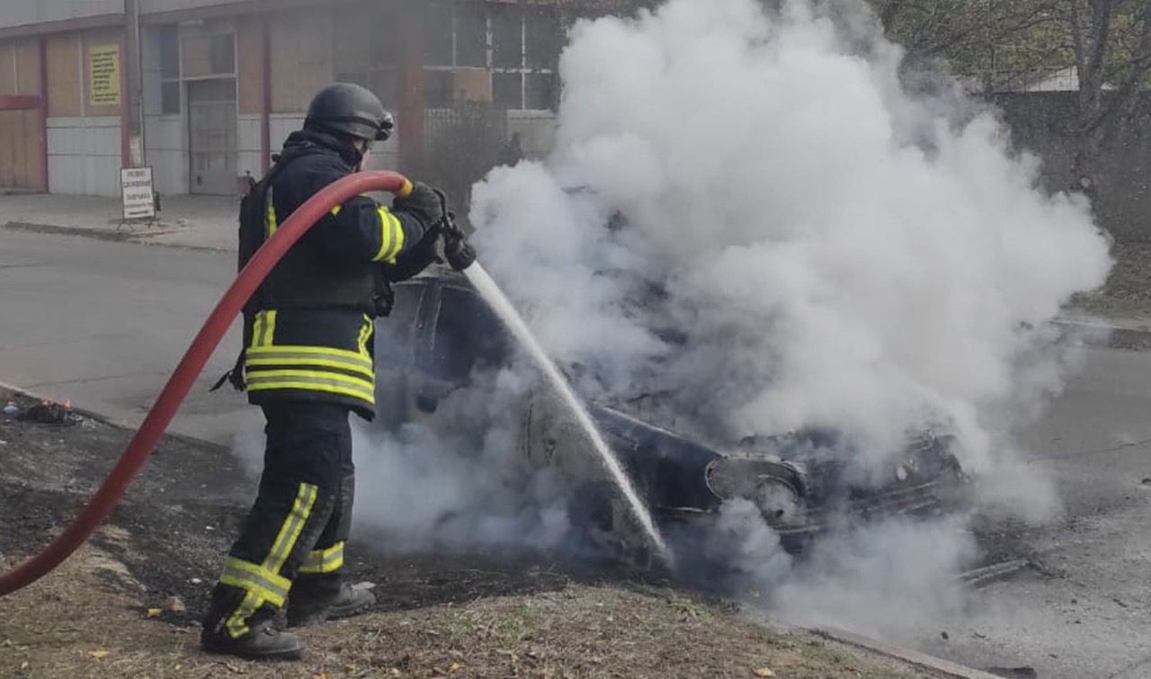 A Ukrainian firefighter in full gear extinguishes a fire on the smoking wreckage of a car following a Russian drone attack