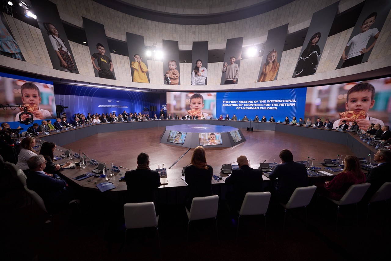 Wide view of a conference room where delegates for the International Coalition for the Return of Ukrainian Children are meeting, with large screens displaying portraits of children