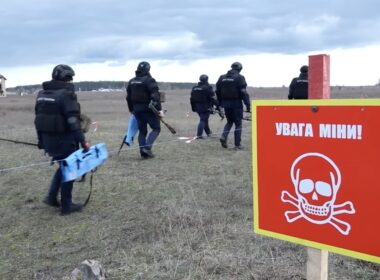 A team of Ukrainian deminers in full gear walks into a field past a red and yellow "Danger Mines!" warning sign with a skull and crossbones