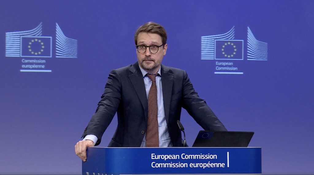 A European Commission spokesperson stands at a podium during a press briefing, discussing new rules for Schengen visas for Russians