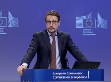 A European Commission spokesperson stands at a podium during a press briefing, discussing new rules for Schengen visas for Russians