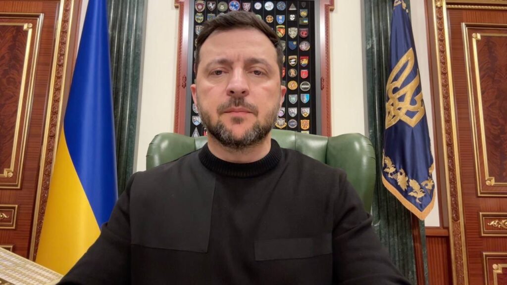 President Volodymyr Zelenskyy sitting in a green leather chair in his office, wearing a black sweatshirt, with the Ukrainian flag and a display of military unit patches visible in the background
