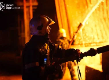 Ukrainian firefighter in helmet holds hose while battling intense orange flames at night, with second rescuer visible in background