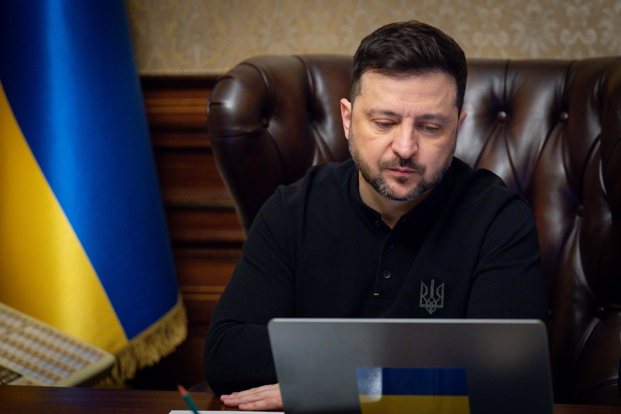 Ukrainian President Volodymyr Zelenskyy at his desk with a laptop, Ukrainian flag visible, during an online press conference