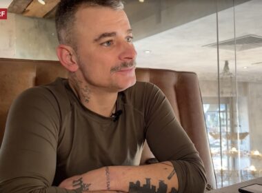 Avi M., a tattooed man in his late 40s with gray hair and mustache, sits in a brown leather booth during an interview with Swiss broadcaster SRF about his service in Ukraine's military