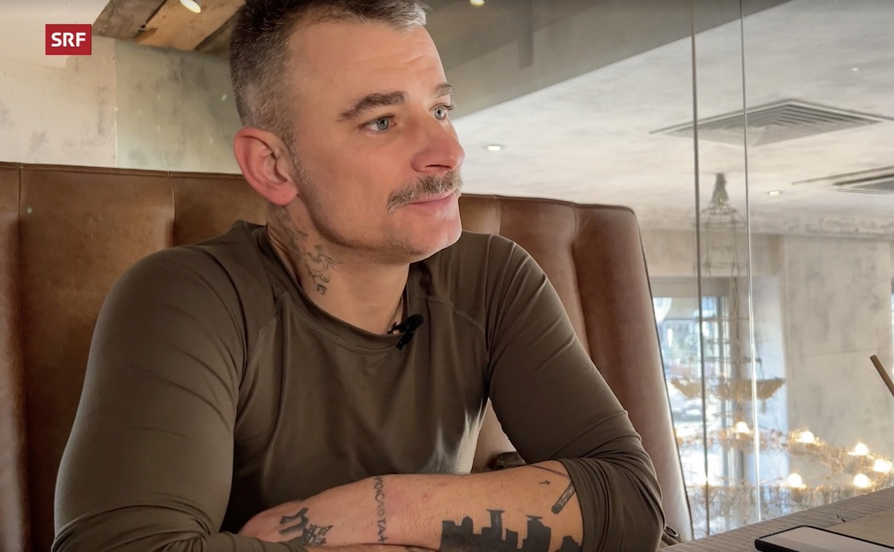 Avi M., a tattooed man in his late 40s with gray hair and mustache, sits in a brown leather booth during an interview with Swiss broadcaster SRF about his service in Ukraine's military