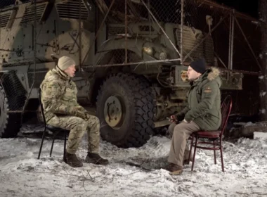 Ukrainian Commander-in-Chief Oleksandr Syrskyi in military uniform sits across from journalist Andriy Drozda in front of a military vehicle with anti-drone cage armor during a snowy winter interview in eastern Ukraine.