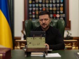 Ukrainian President Volodymyr Zelenskyy sits at his desk during a video call, with Ukrainian flag and military unit patches visible behind him