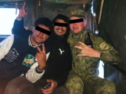 Two migrants with obscured faces pose alongside a Belarusian soldier in camouflage uniform inside a military transport vehicle, all making hand gestures