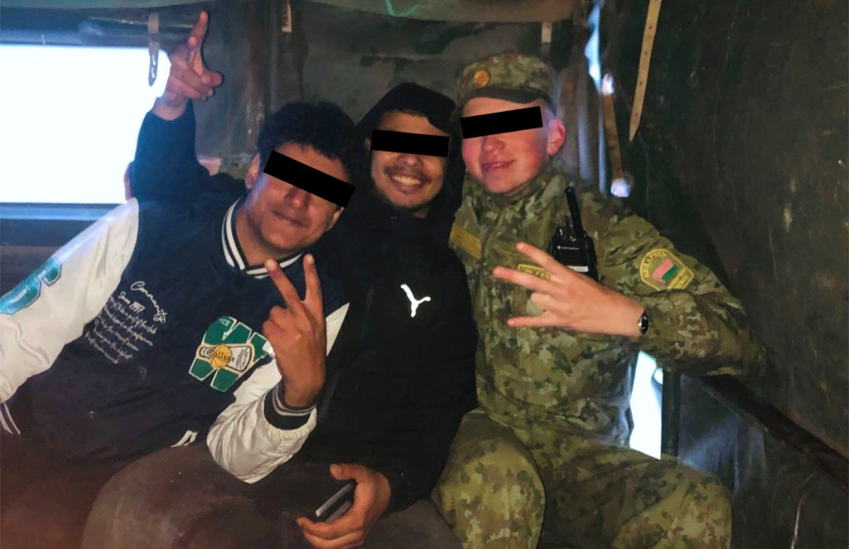 Two migrants with obscured faces pose alongside a Belarusian soldier in camouflage uniform inside a military transport vehicle, all making hand gestures
