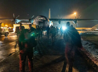 Danish military personnel on a nighttime tarmac in Greenland approach a C-130 Hercules transport aircraft, with snow visible on the ground and aircraft lights illuminating the scene