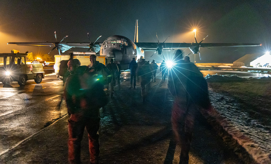 Danish military personnel on a nighttime tarmac in Greenland approach a C-130 Hercules transport aircraft, with snow visible on the ground and aircraft lights illuminating the scene