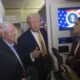 President Donald Trump gestures while speaking to reporters alongside Senator Lindsey Graham aboard Air Force One, with the presidential seal visible on a screen behind them.