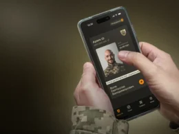 Ukrainian soldier in pixelated camouflage holds smartphone showing Army+ app with digital military ID displaying photo, name, and birth date