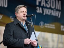 Ukrainian Ambassador to the Czech Republic Vasyl Zvarych speaks at a microphone during a pro-Ukraine rally, wearing a dark coat over a traditional Ukrainian vyshyvanka shirt with a blue-and-yellow ribbon pin.
