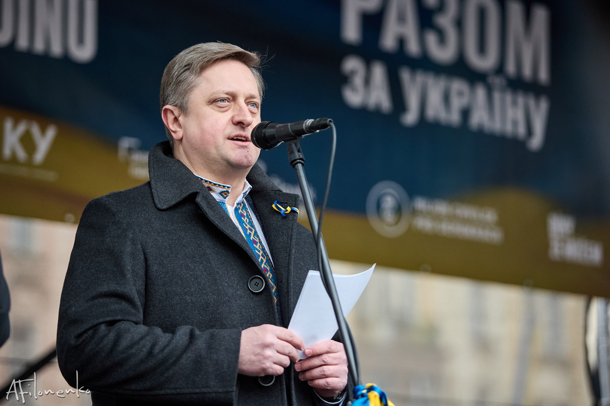 Ukrainian Ambassador to the Czech Republic Vasyl Zvarych speaks at a microphone during a pro-Ukraine rally, wearing a dark coat over a traditional Ukrainian vyshyvanka shirt with a blue-and-yellow ribbon pin.