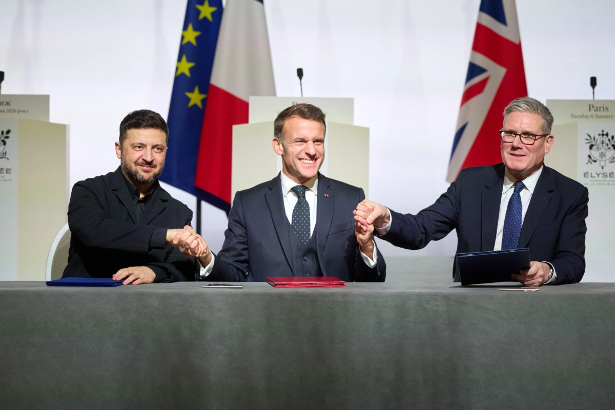 Zelenskyy, Macron, and Starmer seated at signing table, holding raised hands together, with EU, French, and UK flags behind them