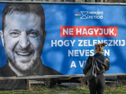 A woman looks up at a Fidesz "Nemzeti Petíció" billboard in Hungary featuring a black-and-white image of Volodymyr Zelenskyy laughing, with the Hungarian text: "Ne hagyjuk, hogy Zelenszkij nevessen a vi[lágon]"—"Don't let Zelenskyy laugh at the [world]."