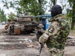 Russian soldier with Z insignia stands near a destroyed armored vehicle on a street in occupied Rubizhne, Luhansk Oblast, where occupation authorities have now manufactured a food shortage by blocking civilian deliveries