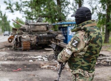 Russian soldier with Z insignia stands near a destroyed armored vehicle on a street in occupied Rubizhne, Luhansk Oblast, where occupation authorities have now manufactured a food shortage by blocking civilian deliveries