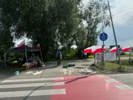 Polish demonstrators under a black canopy with red-and-white Polish flags lining a roadside fence at the Medyka border crossing, with a cyclist passing on the bicycle lane.