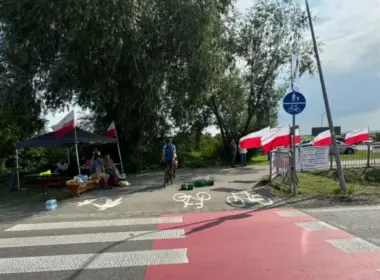 Polish demonstrators under a black canopy with red-and-white Polish flags lining a roadside fence at the Medyka border crossing, with a cyclist passing on the bicycle lane.