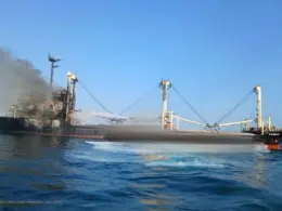 A damaged cargo ship burns at sea as thick black smoke rises from its stern and water sprays across the vessel.