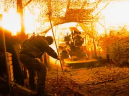 A soldier from Ukraine’s 100th Mechanized Brigade operates an artillery gun firing beneath camouflage netting.