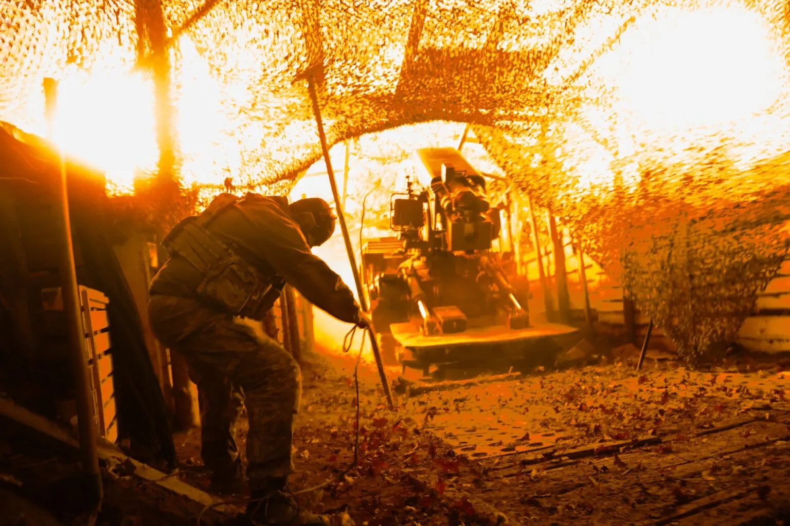 A soldier from Ukraine’s 100th Mechanized Brigade operates an artillery gun firing beneath camouflage netting.