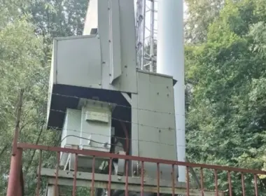 Close-up of equipment at the base of a communications tower behind a metal fence in a wooded area.
