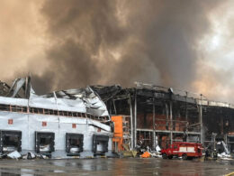 Firefighters beside a heavily damaged warehouse holding Bosch products as smoke rises from its collapsed roof.