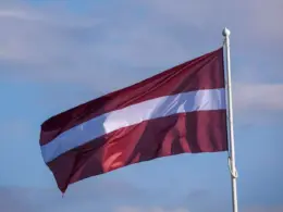 Latvian flag flying from a white flagpole against a blue, lightly clouded sky.