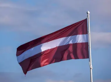 Latvian flag flying from a white flagpole against a blue, lightly clouded sky.