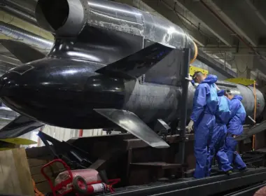 Three workers in blue protective suits inspect a large black Flamingo cruise missile on a factory assembly line.