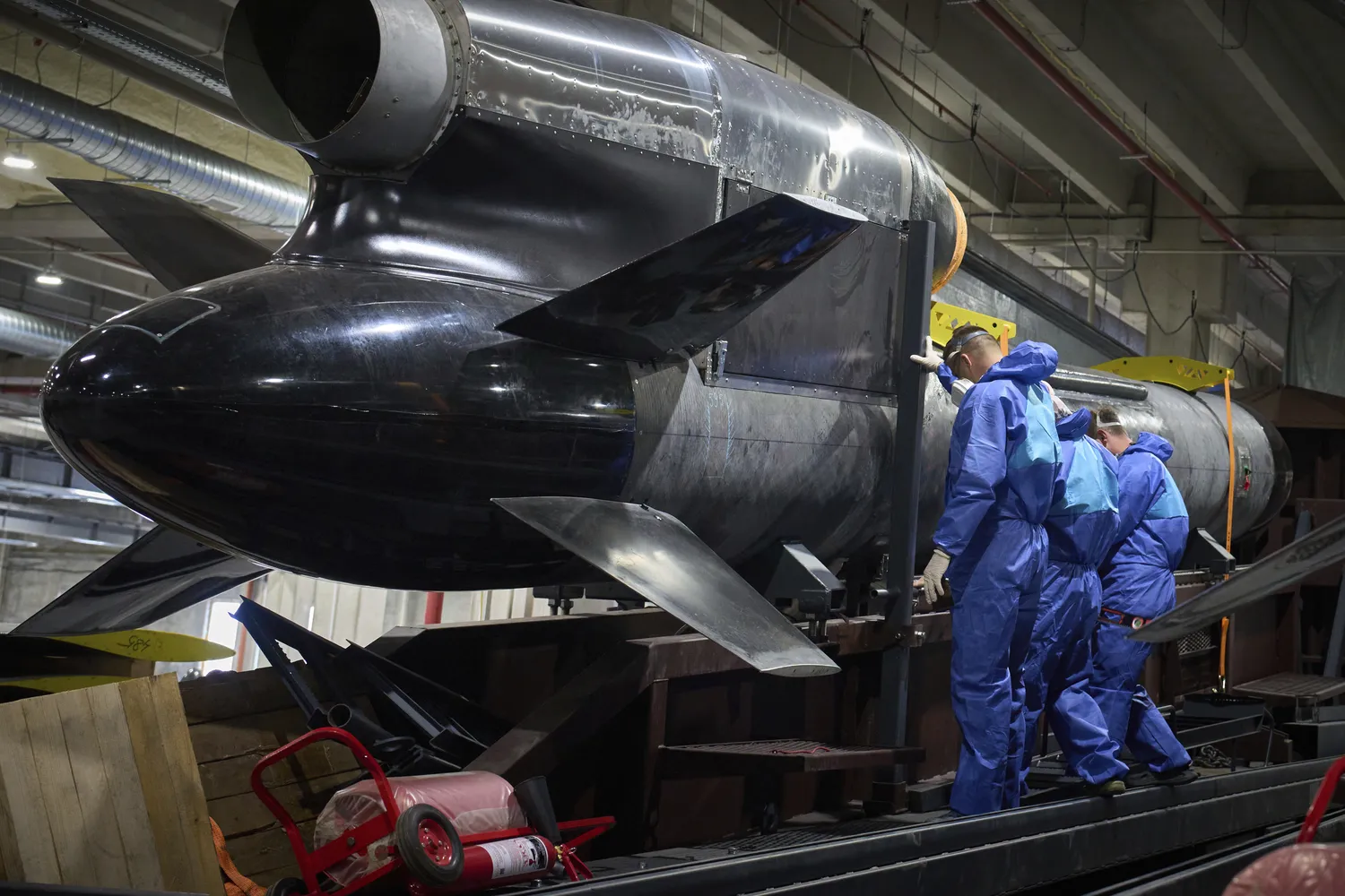 Three workers in blue protective suits inspect a large black Flamingo cruise missile on a factory assembly line.