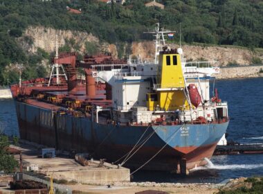 The bulk carrier now named LAMAR S, with SPLIT painted on its stern, moored beside a concrete quay near a wooded coastline.