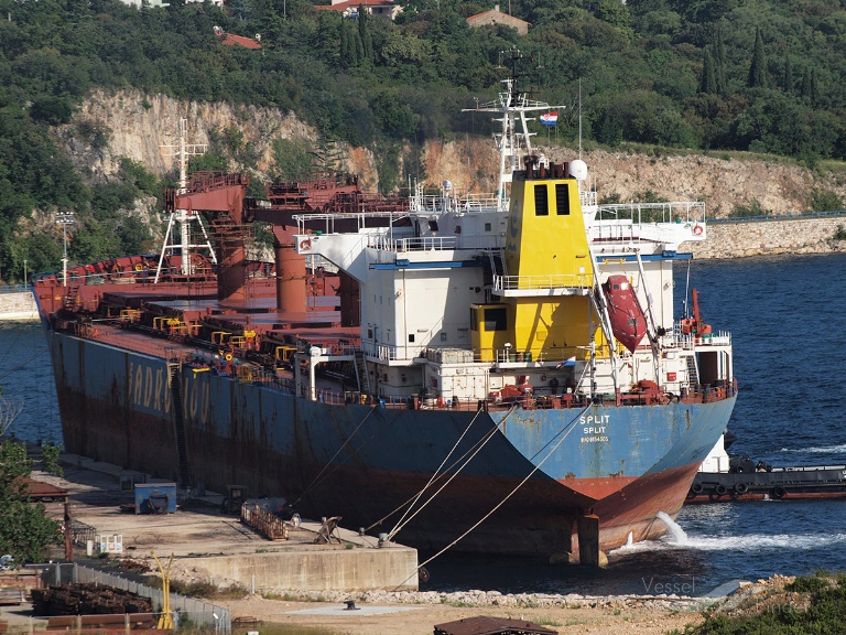 The bulk carrier now named LAMAR S, with SPLIT painted on its stern, moored beside a concrete quay near a wooded coastline.