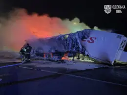 Firefighters stand beside two burned truck trailers covered in ice as smoke and orange firelight fill the night sky.