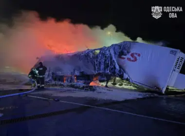 Firefighters stand beside two burned truck trailers covered in ice as smoke and orange firelight fill the night sky.