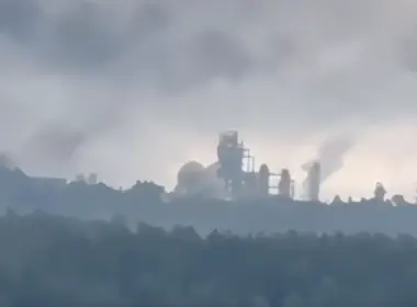 Distant refinery structures silhouetted beneath a cloudy sky, with several gray plumes rising above the industrial complex.