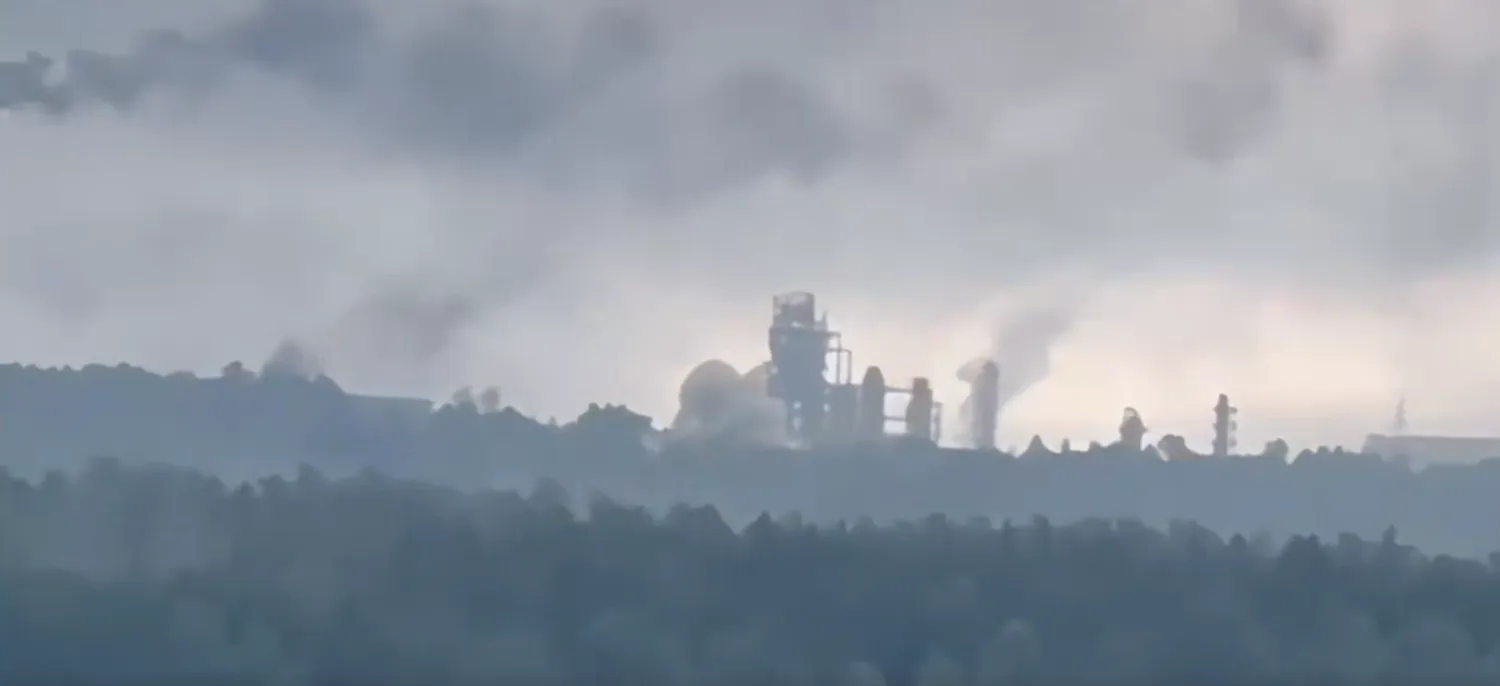 Distant refinery structures silhouetted beneath a cloudy sky, with several gray plumes rising above the industrial complex.
