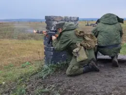 Two soldiers kneel behind a low brick barrier and fire rifles across an open training field.
