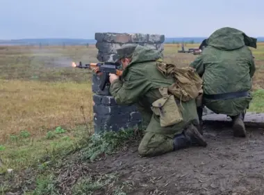Two soldiers kneel behind a low brick barrier and fire rifles across an open training field.