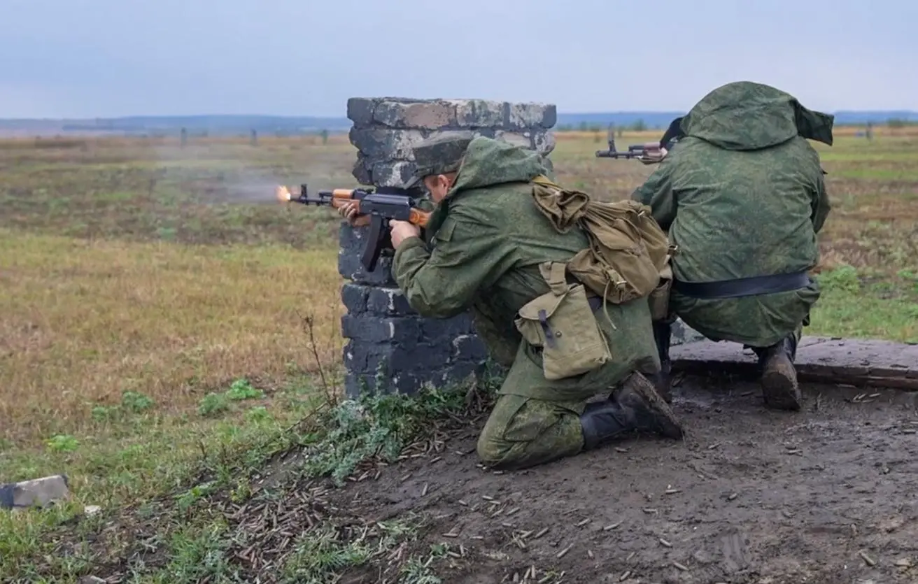 Two soldiers kneel behind a low brick barrier and fire rifles across an open training field.
